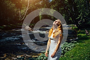 Beautiful woman in white dress with long red hair raised face to the sun on the bank of river in green summer forest.  Portrait of