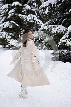 Beautiful woman in white fur posing near spruces in snow. Winter