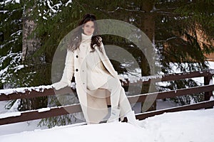 Beautiful woman in white fur posing near spruces in snow. Winter