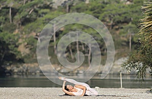 A beautiful woman in a white gymnastic jumpsuit performs a yoga pose while lying down.