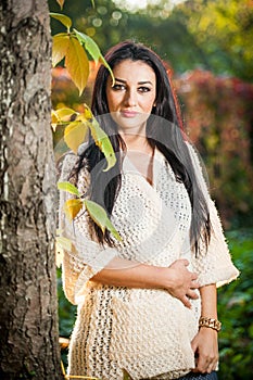 Beautiful woman in white posing in autumnal park. Young brunette woman spending time in autumn near a tree in forest