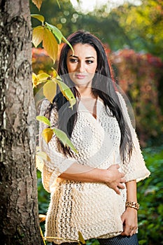 Beautiful woman in white posing in autumnal park. Young brunette woman spending time in autumn near a tree in forest