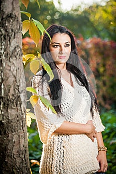 Beautiful woman in white posing in autumnal park. Young brunette woman spending time in autumn near a tree in forest