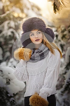 Beautiful woman in white pullover with over-sized fur cap enjoying the winter scenery in forest. Blonde girl posing in winter