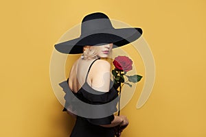 Beautiful woman in wide black broad brim hat with blonde hair and makeup holding red rose flower on yellow background