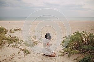 Beautiful woman with windy hair sitting on sandy beach on background of green grass and sea, calm tranquil moment. Stylish young