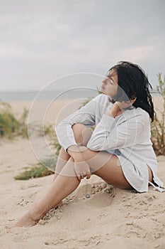 Beautiful woman with windy hair sitting on sandy beach on background of green grass and sea, calm tranquil moment. Stylish young