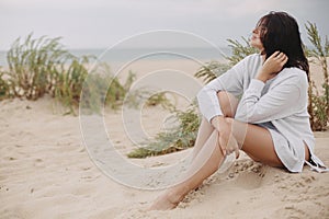 Beautiful woman with windy hair sitting on sandy beach on background of green grass and sea, calm tranquil moment. Stylish young