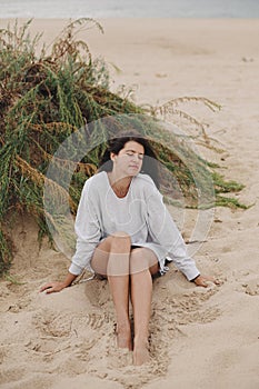Beautiful woman with windy hair and in sweater sitting on sandy beach on background of green grass and sea, calm tranquil moment.