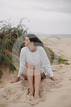 Beautiful woman with windy hair and in sweater sitting on sandy beach on background of green grass and sea, calm tranquil moment.