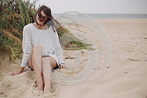 Beautiful woman with windy hair and in sweater sitting on sandy beach on background of green grass and sea, calm tranquil moment.