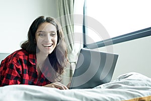Beautiful woman working on a laptop with smiling and lying down on the bed at a condominium in the morning