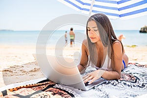 Beautiful woman working online on laptop while lying on beach under sun umbrella near sea. Happy smiling freelancer girl relaxing