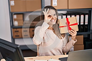 Beautiful woman working at small business ecommerce holding gift smiling happy doing ok sign with hand on eye looking through