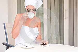 Beautiful woman wrapped in towel after shower with patches under eyes is drinking coffee and using her phone at the hotel terrace