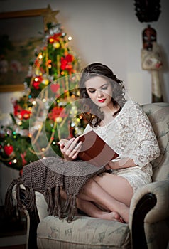 Beautiful woman with Xmas tree in background reading a book sitting on chair. Portrait of a woman reading a book sitting