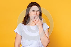 Beautiful woman yawns isolated over yellow background, wears casual white t shirt, has wavy hair, keeps hand near mouth. Free