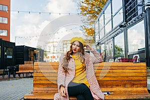 Beautiful woman in yellow beret sits on the street on the bench, depicts emotions. Summer time, yellow colors