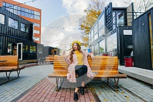 Beautiful woman in yellow beret sits on the street on the bench, depicts emotions. Summer time, yellow colors