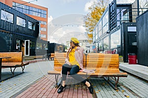 Beautiful woman in yellow beret sits on the street on the bench, depicts emotions. Summer time, yellow colors