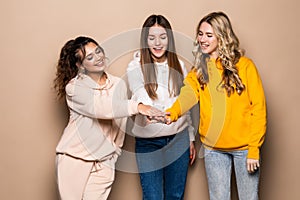 Beautiful three women friends standing with hands on top each other and smiling  on beige background