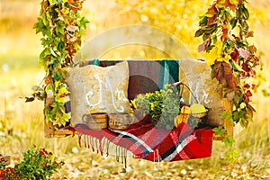 Beautiful wooden swing decorated with a blanket, leaves and flowers, hanging on a tree in autumn garden. Concepts of romance,