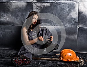 Beautiful worker with a pickaxe and helmet. Seductive and beautiful woman miner sitting on a floor on steel