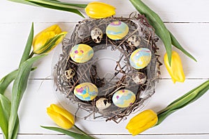 Beautiful yellow tulips with colorful quail and chicken eggs in wreath on white wooden background.