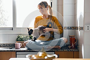 Beautiful youg woman sitting on kitchen table while stroking a cat sitting on her lap at home