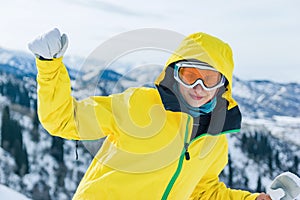 Beautiful youg woman tourist in yellow jacket, ski glasses mask and white mittens on a background of mountains looking