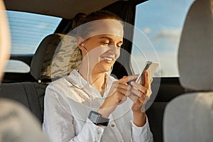 Beautiful young adult caucasian woman wearing white shirt moving in automobile using mobile phone and listening to music on