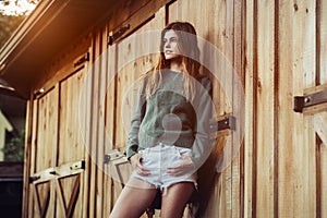 Beautiful young adult country woman posing near barn farm wooden doors at sunset time wearing white shorts and green sweater.