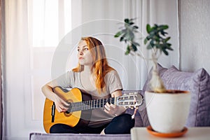 Beautiful young adult girl with long red hair playing the guitar sitting at home in light interior, selective focus. Thoughtful