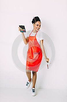 Beautiful young African American beautician woman holding set of make up brushes and eye-shadows, isolated on white