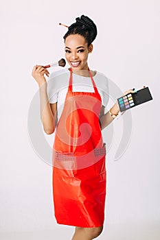 Beautiful young African American beautician woman holding set of make up brushes and eye-shadows, isolated on white