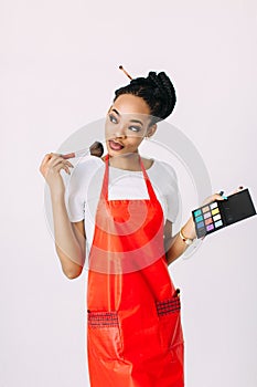 Beautiful young African American beautician woman holding set of make up brushes and eye-shadows, isolated on white