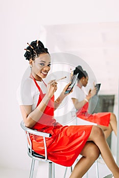 Beautiful young African American beautician woman holding set of make up brushes and watching in the mirror