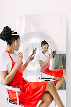 Beautiful young African American beautician woman holding set of make up brushes and watching in the mirror