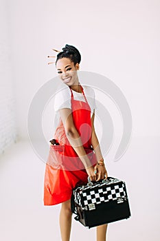 Beautiful young African American beautician woman holding set of makeup equipment in special case, isolated on white