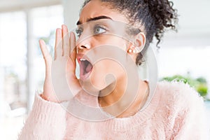 Beautiful young african american woman with afro hair shouting and screaming loud to side with hand on mouth