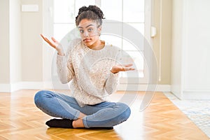 Beautiful young african american woman with afro hair sitting on the floor clueless and confused expression with arms and hands