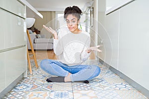 Beautiful young african american woman with afro hair sitting on the floor clueless and confused expression with arms and hands