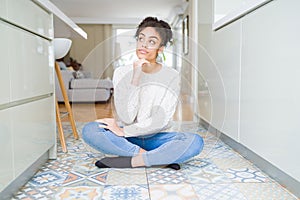 Beautiful young african american woman with afro hair sitting on the floor with hand on chin thinking about question, pensive