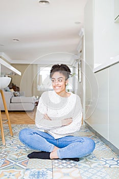 Beautiful young african american woman with afro hair sitting on the floor smiling looking side and staring away thinking