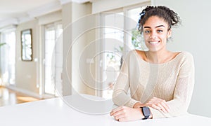 Beautiful young african american woman with afro hair sitting on table at home with a happy and cool smile on face