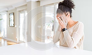 Beautiful young african american woman with afro hair sitting on table at home with sad expression covering face with hands while