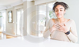 Beautiful young african american woman with afro hair sitting on table at home smiling with hands on chest with closed eyes and