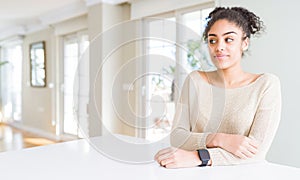 Beautiful young african american woman with afro hair sitting on table at home smiling looking side and staring away thinking