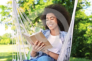 Beautiful young African-American woman reading book in hammock outdoors