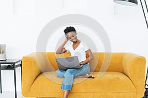 Beautiful young African American woman in white t-shirt and blue jeans using laptop sitting on yellow sofa in bright modern